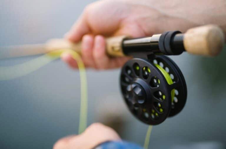 A close-up of a hand holding a fly fishing rod and reel, with the line pulled taut, showcasing the fine craftsmanship and focus needed for fly fishing. The background is softly blurred, emphasizing the equipment and technique.