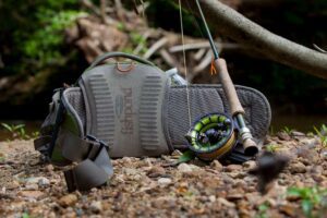 A fly fishing rod and gear pack resting on the riverbank, surrounded by natural greenery, showcasing the essential tools needed for a successful fishing trip.