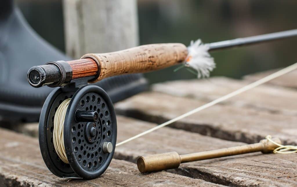 A of what what is the best weight for backing fly fishing looks like , with a fly fishing rod with a cork handle and a black fly reel, resting on a rustic wooden surface. A pair of wading boots is partially visible in the background, suggesting a fly fishing setup ready for use.