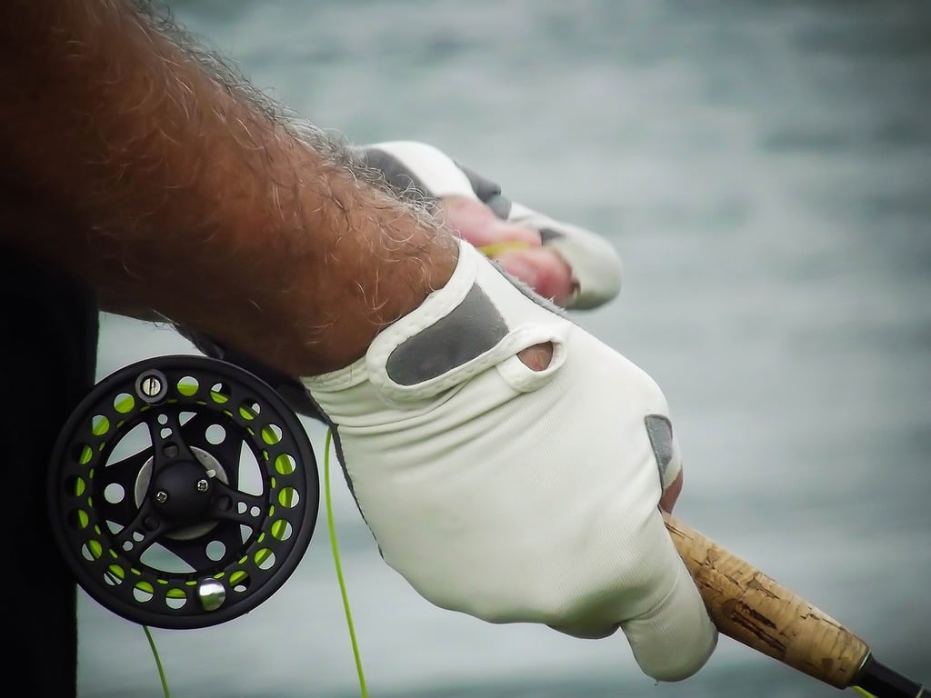 A close-up of a fly fisherman's hands, wearing gloves, gripping a fly fishing rod with a cork handle. The reel is visible, wound with green line, against a blurred background of water, highlighting the precise and attentive nature of fly fishing.